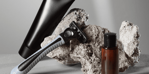 A razor, black aftershave tube, and brown cologne bottle are artfully arranged on rugged stone, showcasing essential grooming products for aftershave vs cologne.