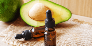 Two small dropper bottles filled with avocado oil for beard care are placed on a rustic cloth next to a fresh avocado cut in half, revealing its green flesh and large seed, set on a wooden table.