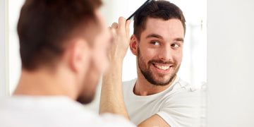 Smiling man styling his hair with a comb in front of a mirror, contemplating the difference between pomade vs gel for his grooming routine.