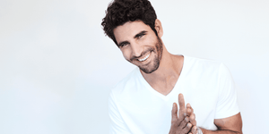 A cheerful man with a 5mm length beard, styled and well-groomed. He has curly dark hair, a bright smile, and is wearing a plain white V-neck t-shirt. The background is light and minimal, focusing the attention on his friendly expression.