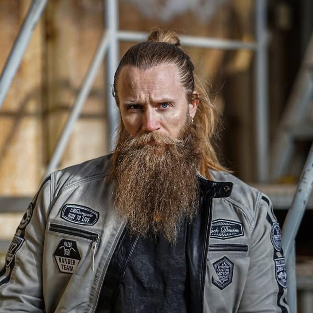 A striking portrait of a man showcasing one of the best beard styles of all time: the Viking Beard. The long, thick, and naturally textured beard exudes rugged masculinity and strength, perfectly complemented by his tied-back long hair. The beard flows freely, giving off a raw and untamed look, while his leather jacket adorned with patches enhances the Norse warrior aesthetic. This powerful and commanding style is ideal for men seeking a bold, timeless, and iconic beard look. The overall appearance captures the essence of the Viking spirit, blending tradition with modern edge.