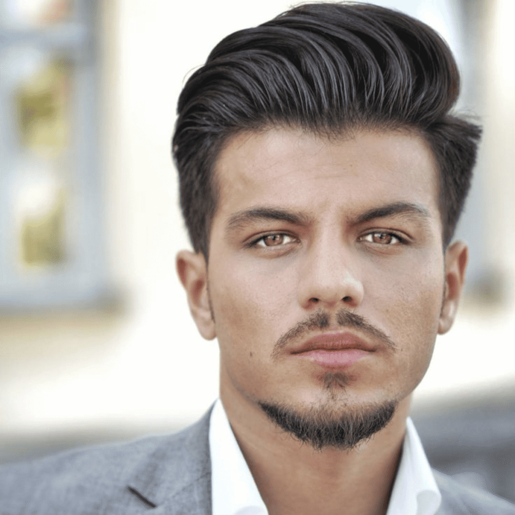 A portrait of a stylish young man showcasing one of the best beard styles, the Dapper Goatee. He has a neatly trimmed chin beard and a distinct, separate mustache, both perfectly groomed to accentuate his chiseled jawline and pronounced chin. His hair is styled in a sleek, swept-back manner, complementing the sharpness of his facial hair. The background is softly blurred, putting the focus on his confident gaze and the precise detailing of his goatee, epitomizing modern elegance and grooming sophistication.