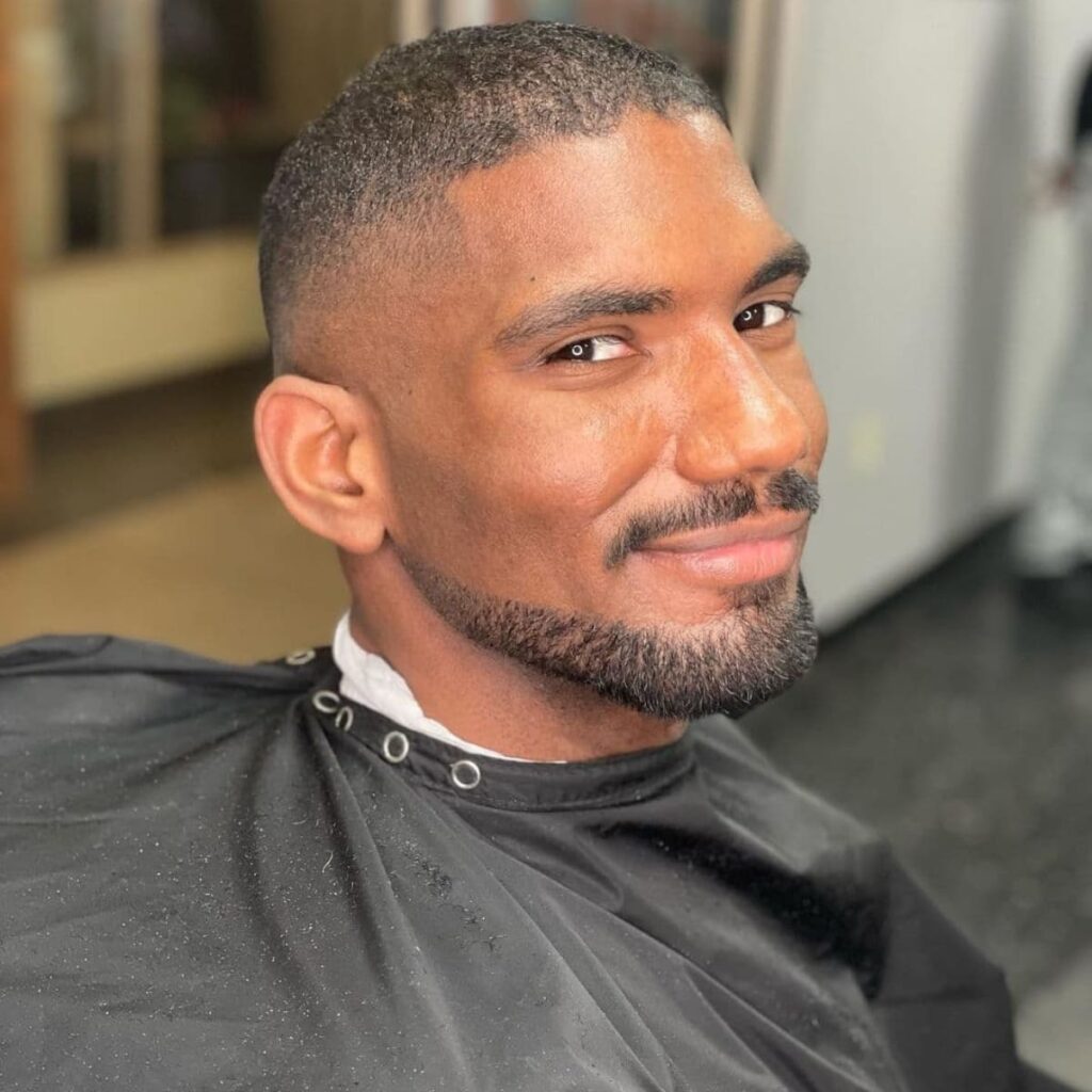A cheerful man in a barber's cape, fresh from a haircut and facial hair trim, smiles warmly at the camera. His skin is smooth and his buzz cut is crisp, enhancing his clean-shaven jawline and neatly styled facial hair that outlines his chin and upper lip. His dark eyes are bright with a spark of joy, complementing his inviting smile. The salon's interior reflects off the cape, suggesting a casual, comfortable grooming session. This image portrays a moment of simple pleasure, capturing the man's polished appearance and content demeanor.