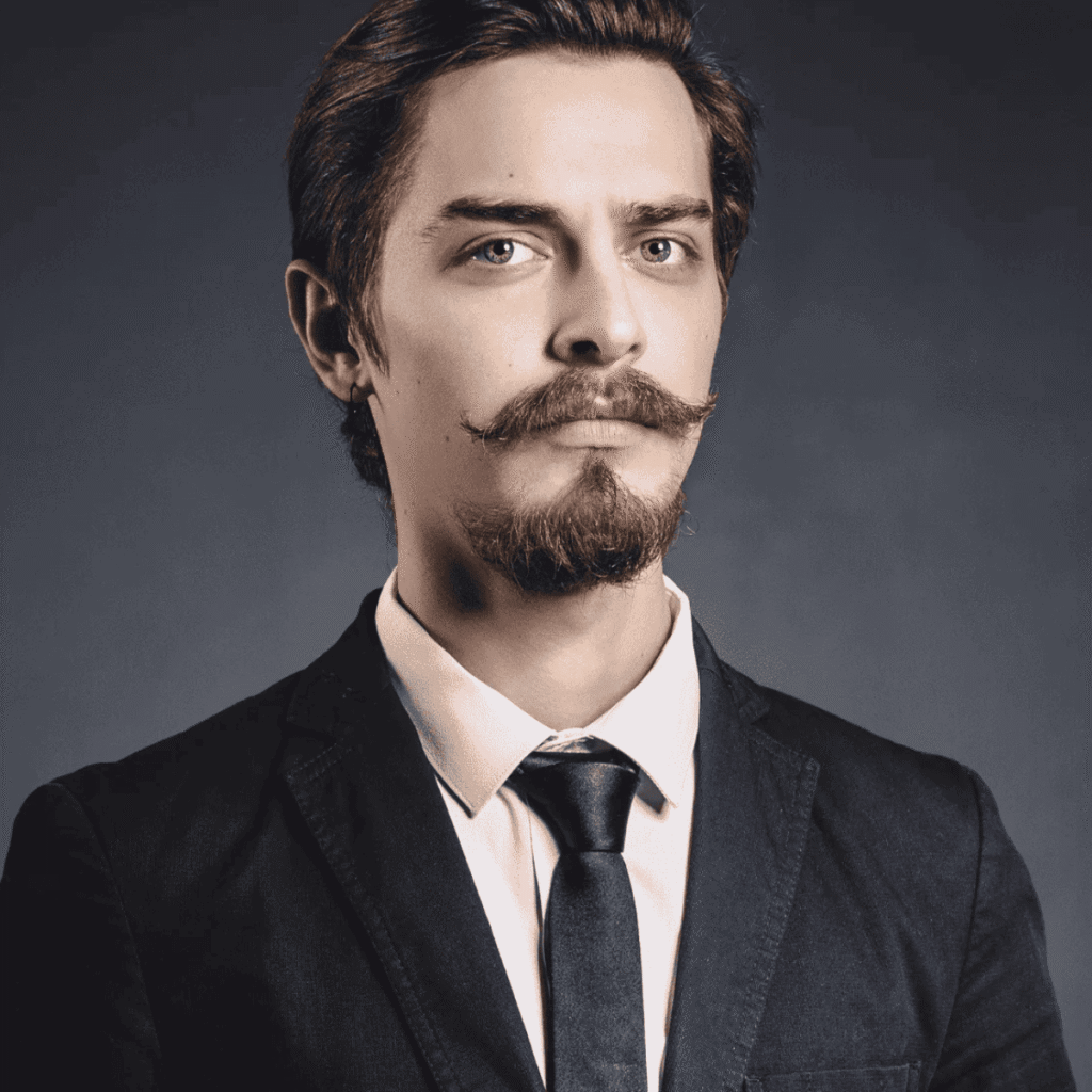 A poised young man dressed in a sophisticated black suit and tie, showcasing a facial hair. His facial hair is meticulously sculpted to highlight a prominent, curled mustache and a pointed chin facial hair, styled to emphasize the angular features of his face. His hair is neatly groomed in a classic, parted style, complementing the vintage look of his facial hair. The subtle gray background enhances the timeless elegance of his appearance, capturing a blend of classic and contemporary grooming excellence.