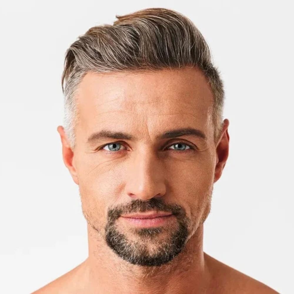 A close-up image of a man with a well-maintained facial hairstyle, featuring a connected mustache and goatee that form a rounded shape around his mouth. His salt-and-pepper facial hair complements his mature yet youthful appearance. The hairstyle is styled neatly with a side-swept look, adding sophistication to his overall presentation. The clean white background highlights his grooming and sharp facial features, emphasizing a polished and professional style.
