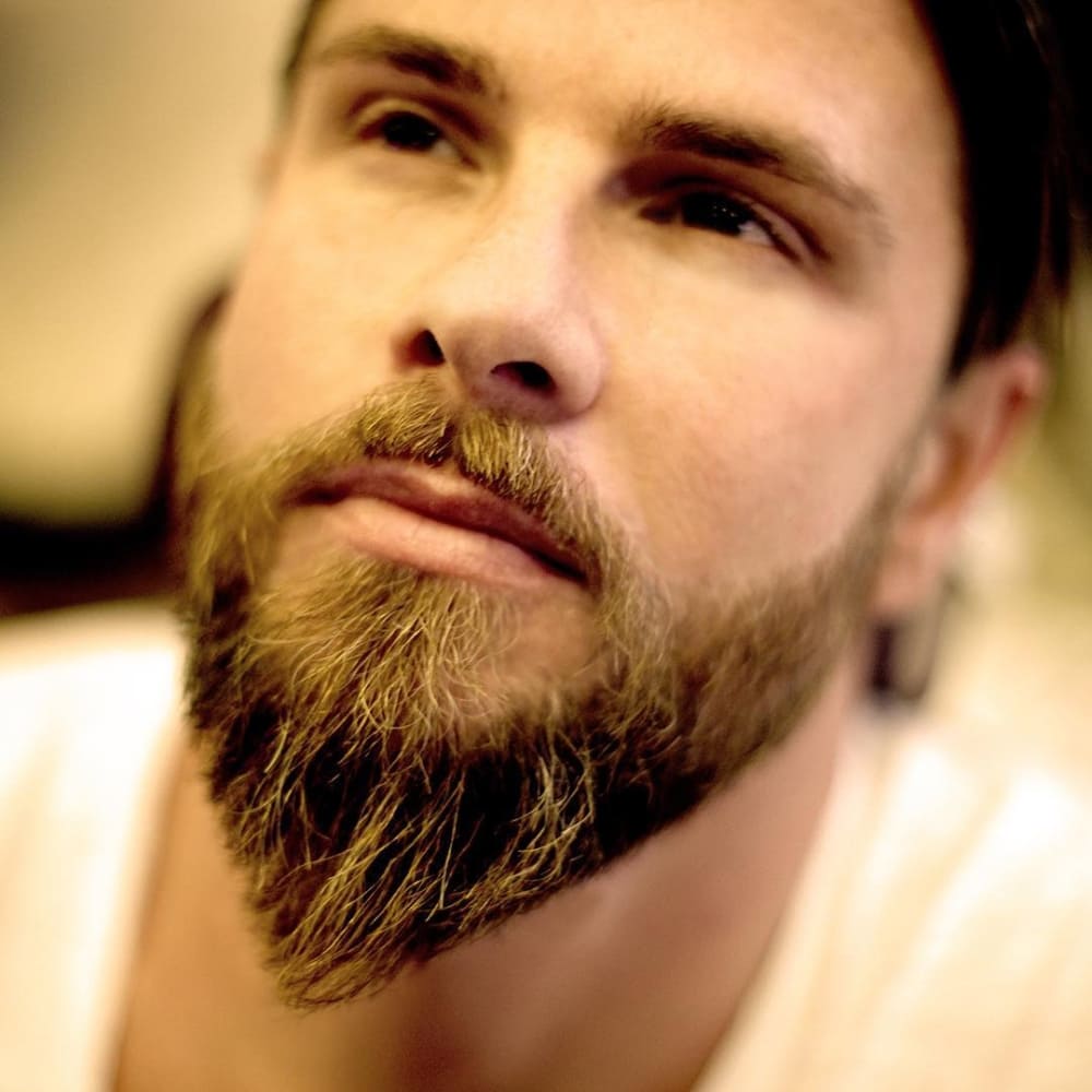 A close-up image of a man showcasing the ducktail beard, one of the timeless beard styles known for its distinctive shape and sophistication. The beard is full and tapered, with longer, pointed growth at the chin, creating a sleek and balanced appearance. The well-groomed facial hair frames the jawline, enhancing the man’s masculine features. His soft, thoughtful expression and slightly blurred background contribute to the focus on the beard’s texture and style. This image perfectly highlights the elegance and versatility of timeless beard styles like the ducktail.