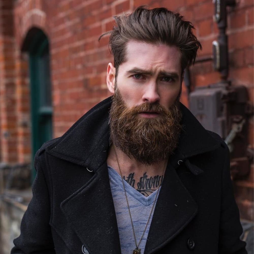 A ruggedly stylish man showcasing the Garibaldi beard, one of the classic beard styles that balances boldness with simplicity. The beard is full and rounded, approximately six to eight inches in length, with a slightly untamed finish that exudes a natural, effortless vibe. His windswept hairstyle complements the beard's rugged aesthetic, while his layered outfit, featuring a dark coat and casual shirt, adds to his approachable yet confident look. The urban brick wall background enhances the contrast of his polished yet natural appearance, making this a striking example of timeless and classic beard styles.