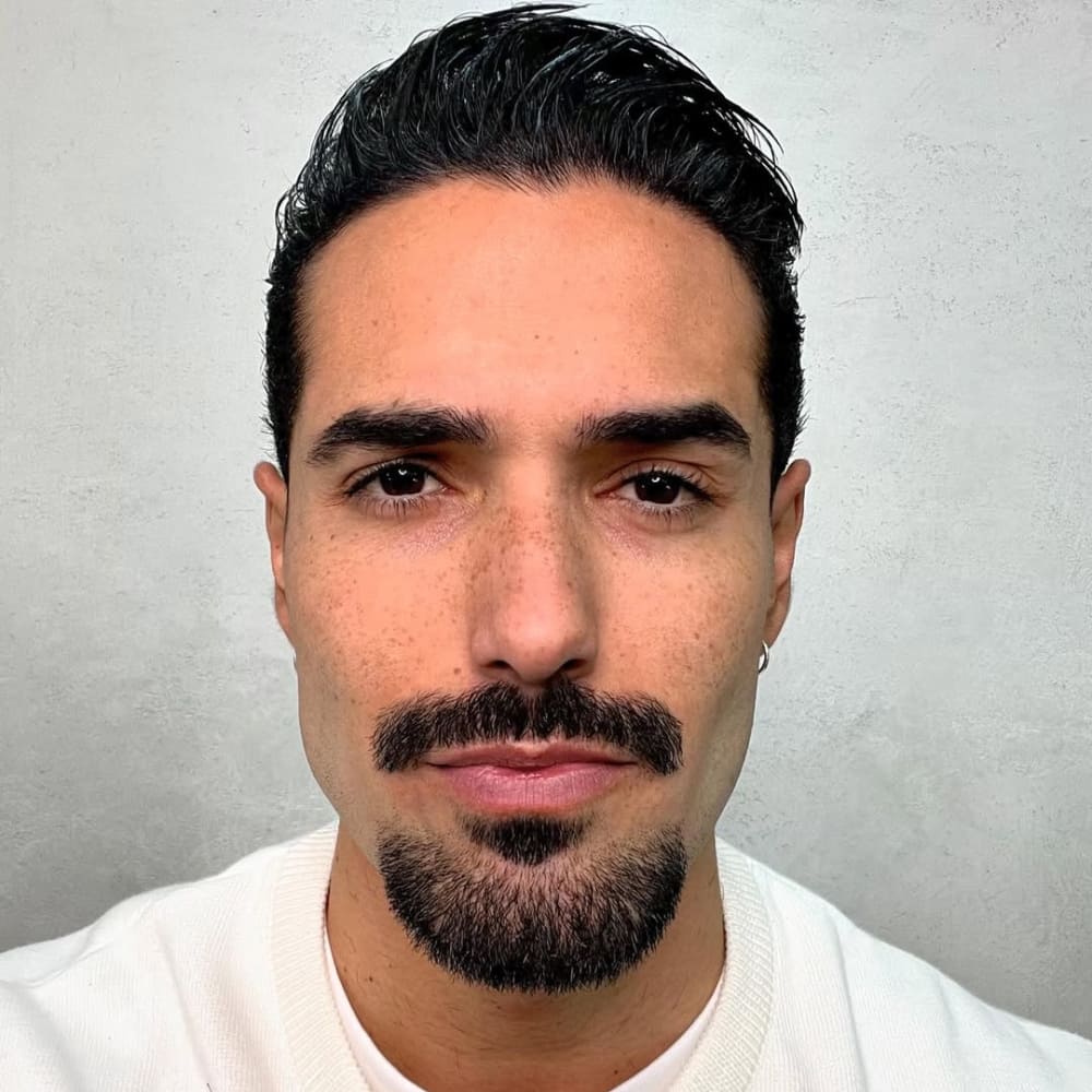 A close-up image of a man with a neatly groomed disconnected goatee beard. The style features a precise patch of facial hair on the chin paired with a sharp, defined mustache, both intentionally separated for a modern, clean look. His smooth, clean-shaven cheeks and jawline emphasize the contrast of the facial hair. The man has well-defined facial features, including a strong chin and symmetrical proportions. His dark, slicked-back hair adds to the polished appearance, and the neutral background highlights the detail and grooming of the goatee. Perfect for showcasing a minimalist and contemporary beard style.