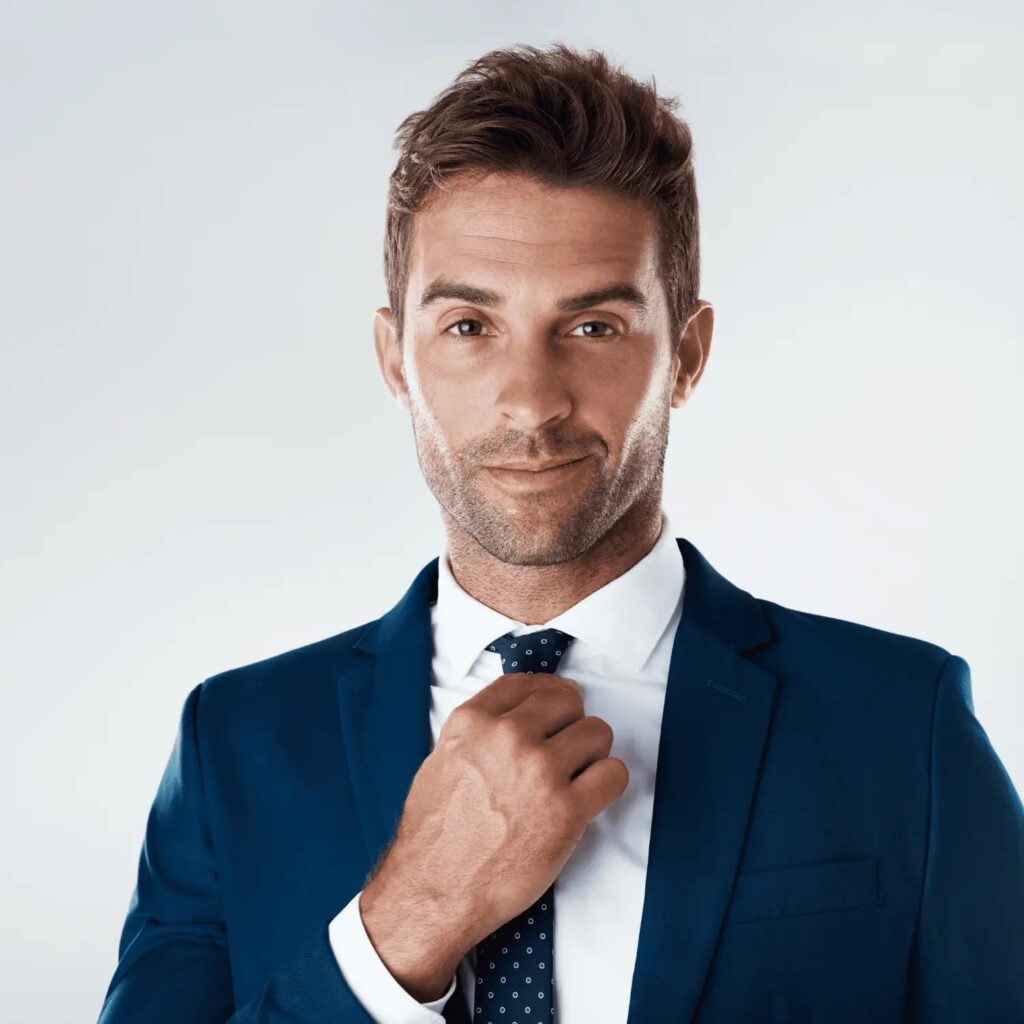 A confident man wearing a tailored navy blue suit and white shirt, adjusting his dark patterned tie. He has a well-groomed stubble beard style, complementing his clean and professional look. The light background enhances the sharpness of his attire and the neat grooming of his beard, ideal for showcasing trendy stubble beard styles.
