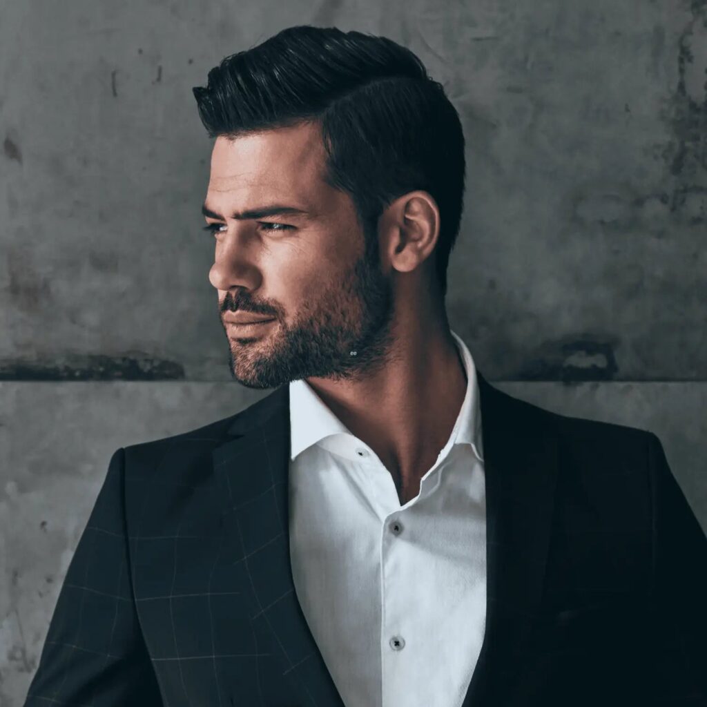 A stylish man in a black checkered suit and crisp white shirt gazes to the side with a confident expression. His neatly combed side-part hairstyle complements his well-maintained stubble beard style, adding a touch of sophistication and masculinity. The concrete-textured background emphasizes the modern and edgy vibe of his look, making it an ideal example of sharp stubble beard styles paired with formal wear.