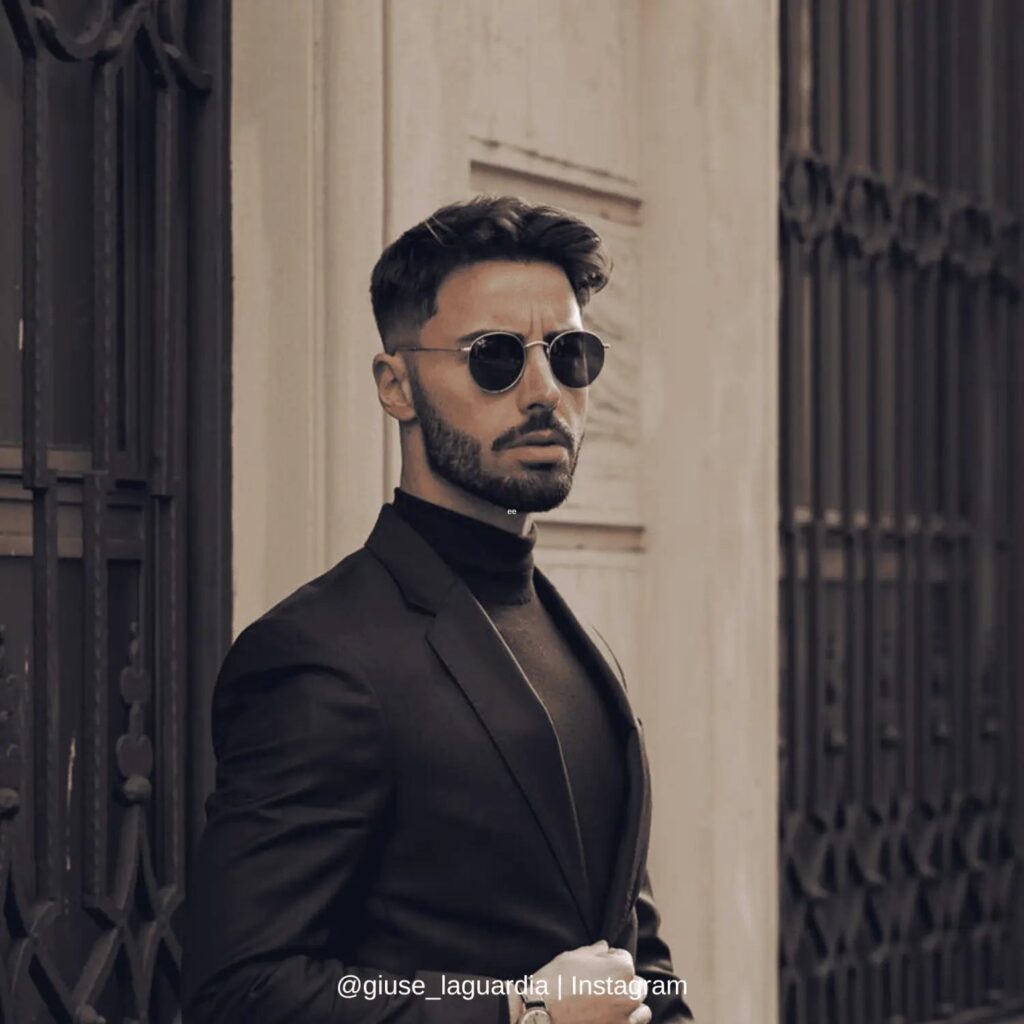 A fashionable man wearing a black suit and turtleneck leans against an ornate iron gate, exuding sophistication and elegance. He sports round sunglasses and a sleek hairstyle, enhancing his confident and modern appearance. The neutral-toned background adds a timeless and classic atmosphere to the composition.