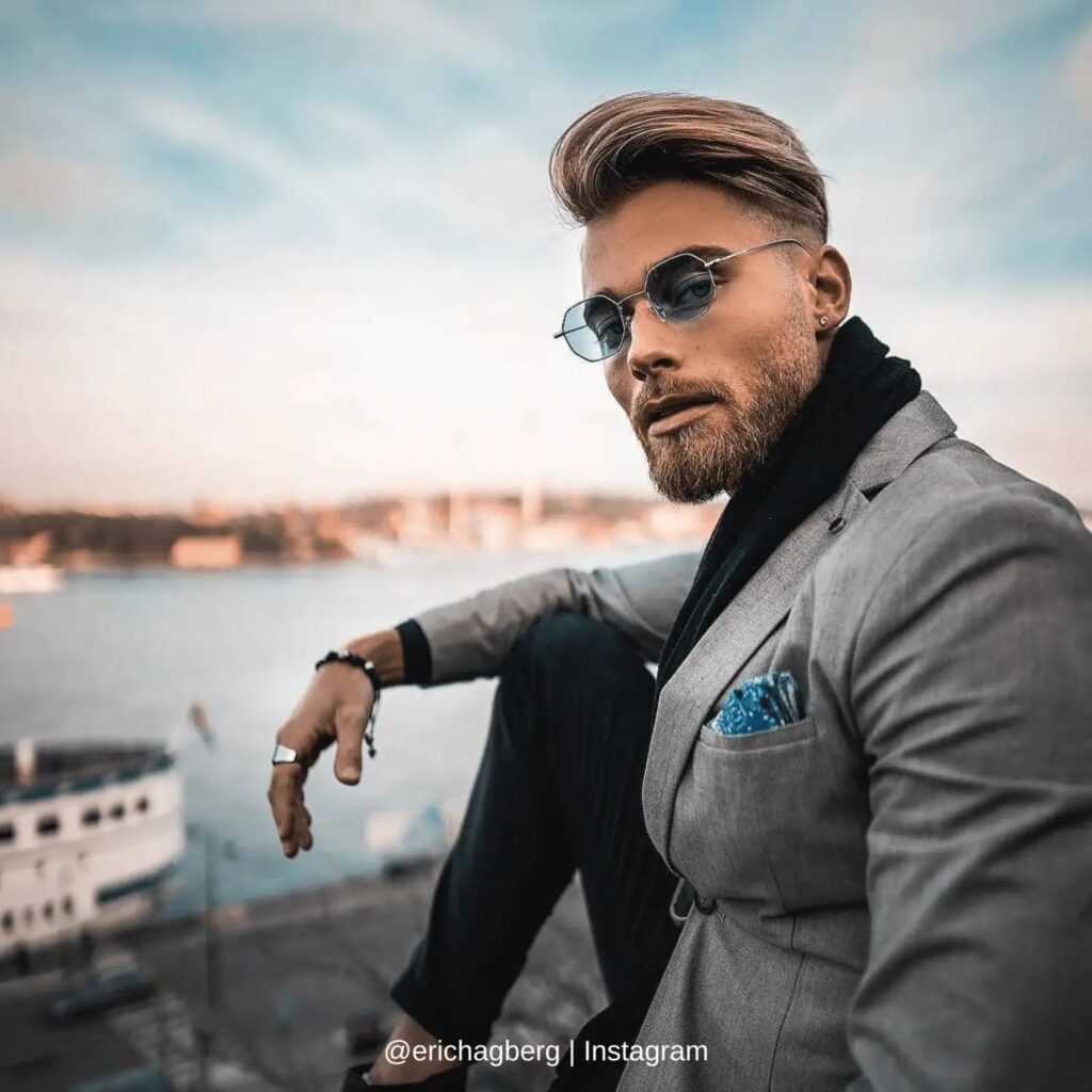 A stylish man sits by a waterfront, wearing a light gray blazer paired with a black scarf and a blue pocket square. He sports round sunglasses, a sleek undercut hairstyle, and a meticulously groomed stubble beard style, exuding a blend of modern elegance and casual confidence. The scenic background of boats and a soft blue sky adds a sophisticated yet relaxed vibe to the image, highlighting trendy stubble beard styles in a fashionable urban setting.