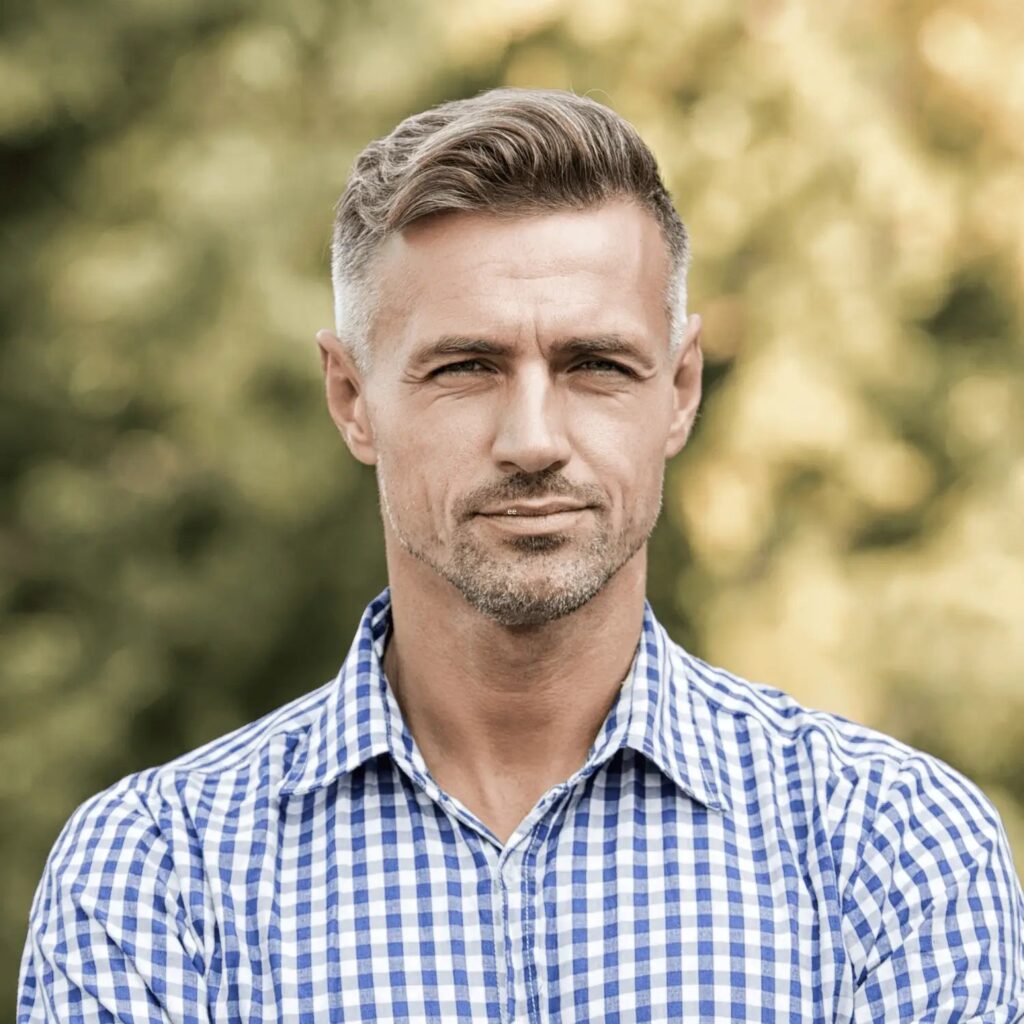 A confident man with a sharp, clean look stands outdoors, wearing a blue and white checkered shirt. His neatly styled hair features a subtle fade, and his expression is composed and self-assured. The blurred green background of trees creates a natural and serene setting, enhancing the focus on his well-groomed appearance.