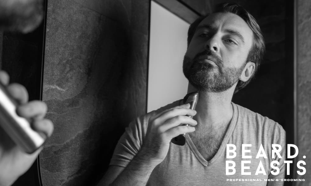 Man shaping his short beard with an electric trimmer in front of a mirror, focusing on clean neckline definition and facial structure.