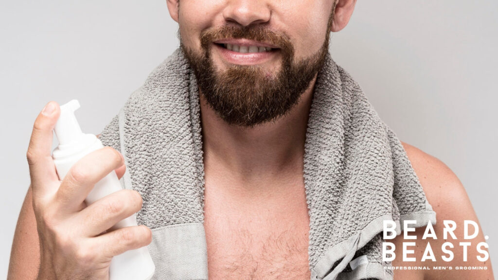 A smiling bearded man with a neatly groomed beard holding a pump bottle of beard wash. A gray towel is draped over his shoulders, suggesting a grooming routine focused on cleanliness and proper beard care. The Beard Beasts logo in the corner reinforces the emphasis on professional men's grooming and maintaining a healthy beard.