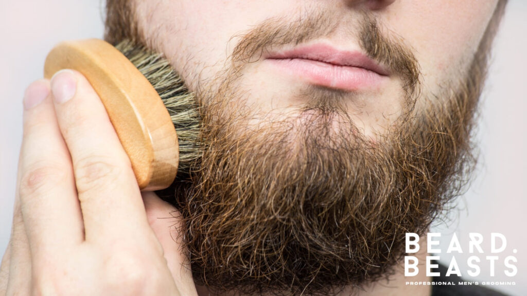A close-up image of a man using a wooden beard brush with natural bristles to groom his full, thick beard. This demonstrates proper beard care, emphasizing the importance of brushing to maintain a healthy, neat, and well-conditioned beard. The Beard Beasts logo in the corner highlights the focus on professional men's grooming.