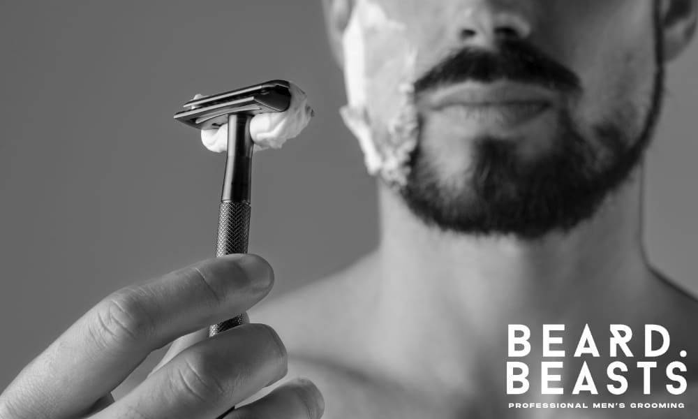 Close-up of a man holding a shaving razor with shaving cream on his cheeks, highlighting the importance of maintaining a clean cheek line even with patchy beard growth.