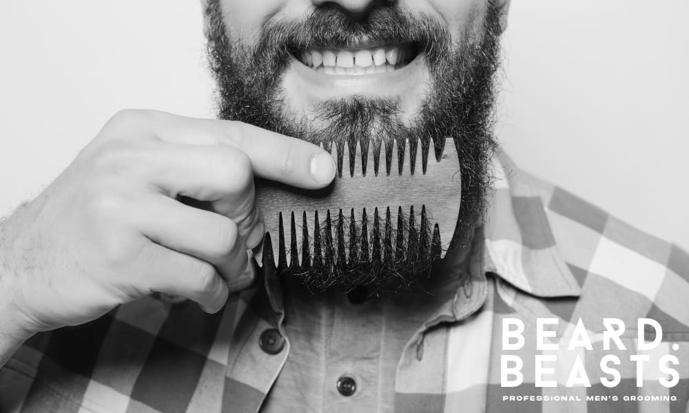 Close-up of a man combing his thick beard with a wooden comb, demonstrating the proper technique for maintaining a soft and healthy beard.