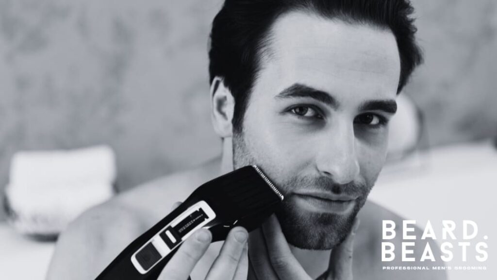 Man using an electric trimmer to groom his facial hair, maintaining a clean and precise look. The setting features a soft, indoor background, emphasizing a professional and effortless grooming routine. His focused expression highlights the importance of proper tools for achieving a well-maintained appearance.