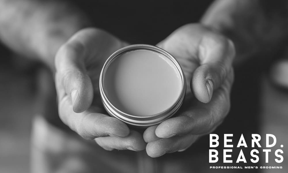 Tattooed hands holding an open tin of beard balm, highlighting product texture and preparation before proper application.