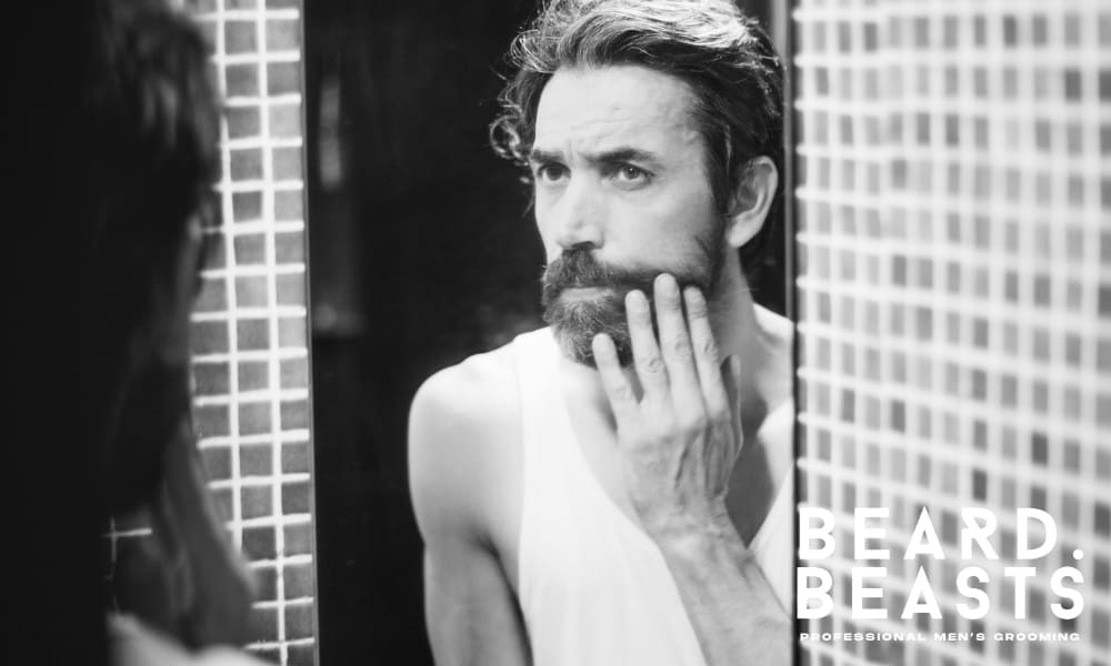 Black and white image of a bearded man looking in the mirror and touching his beard, showing concern about beard pain. The image captures the question, 'why does my beard hurt,' and emphasizes the importance of proper grooming and beard care.