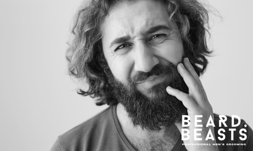 lack and white image of a man with a full beard and long hair, visibly uncomfortable and touching his face, highlighting the topic of 'why does my beard hurt' and addressing common causes of beard pain.