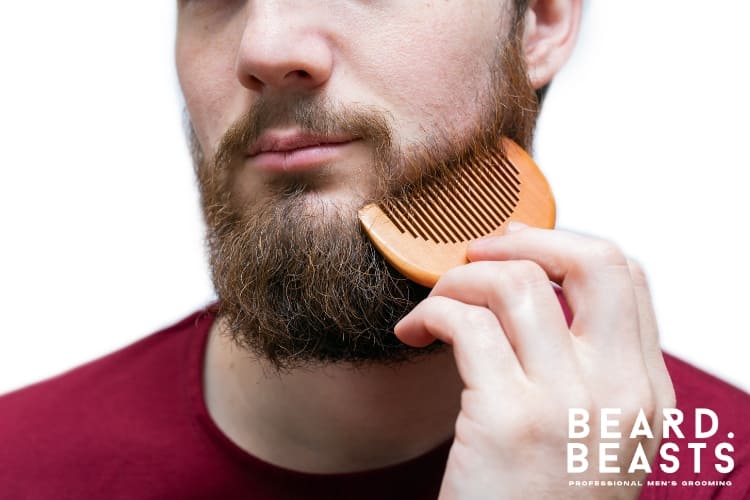 A bearded man grooming his beard using a wooden beard comb, emphasizing proper beard care and maintenance. The comb is held close to the face, showcasing its smooth teeth and natural finish. This highlights the importance of knowing how to clean a beard comb for hygiene and longevity, especially for wooden combs.
