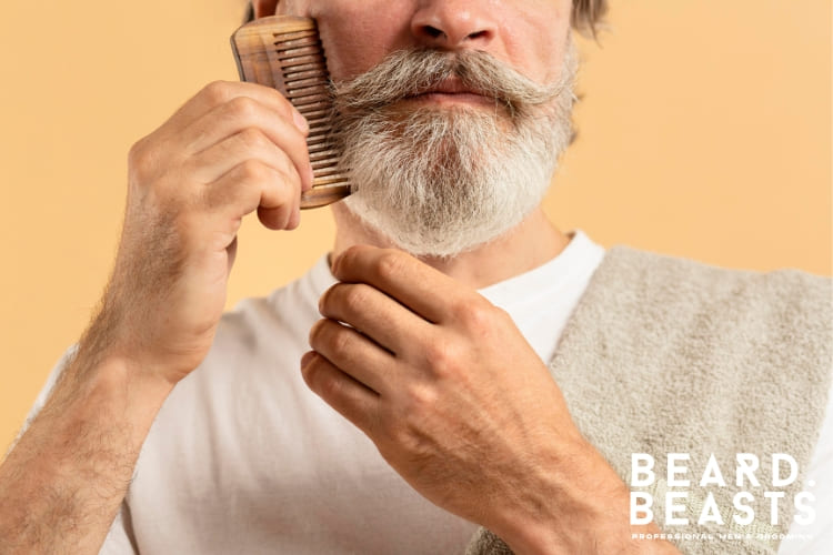A mature man with a well-groomed gray beard and mustache styling his facial hair with a wooden grooming tool. The focus is on maintaining a polished beard, emphasizing the importance of beard care and hygiene. The image aligns with tips on how to clean a beard comb to ensure long-term grooming effectiveness.