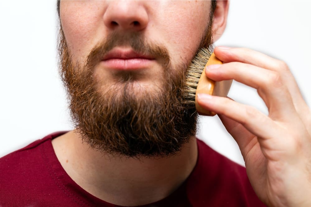 A close-up of a bearded man brushing his beard with a boar bristle brush, demonstrating proper grooming techniques. The image highlights the benefits of brushing your beard, such as smoothing out tangles, exfoliating the skin beneath, and achieving a polished, well-maintained appearance.
