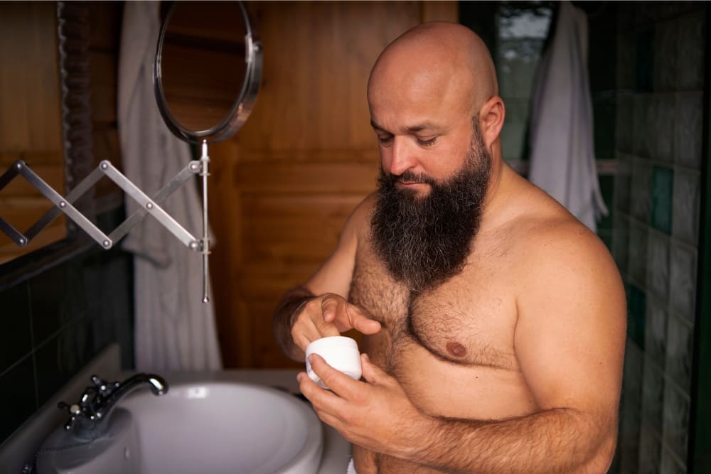 A bald man with a full, well-groomed beard stands in a bathroom, preparing to apply beard balm from a container. The image emphasizes the benefits of brushing your beard, including enhancing beard texture, promoting even product application, and maintaining a healthy, polished look.
