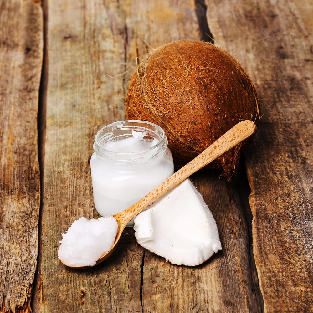 A whole coconut, a glass jar of solidified coconut oil, a wooden spoon holding a scoop of coconut oil, and a piece of fresh coconut flesh, all placed on a rustic wooden surface.