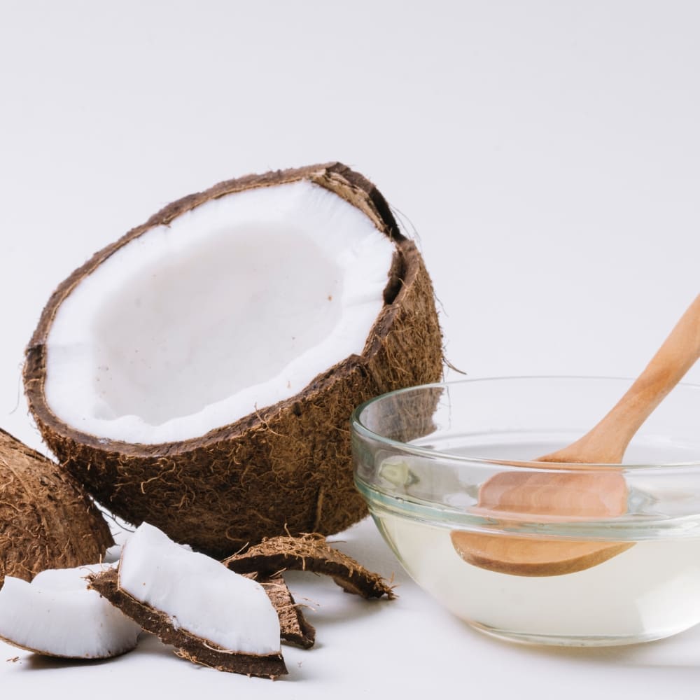 Half of a cracked coconut and small pieces of coconut shell alongside a glass bowl filled with clear coconut oil, with a wooden spoon resting in the bowl, all set against a clean white background.