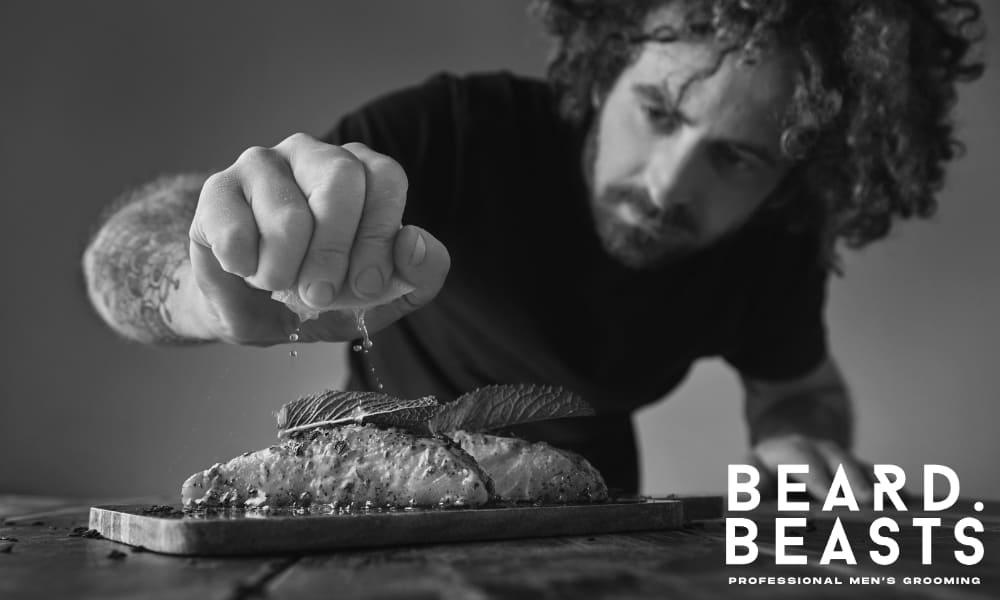 Bearded man preparing salmon with herbs and lemon—cooking nutrient-rich food to support facial hair growth naturally.