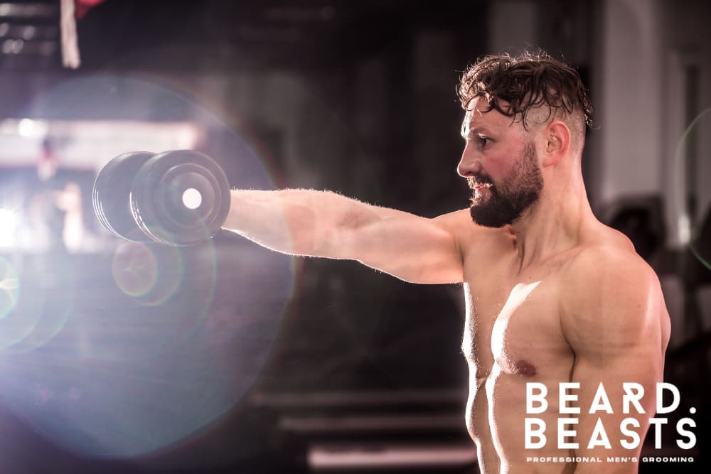 A shirtless man performing strength training with a dumbbell in a gym setting, emphasizing his dedication to fitness and health. The lighting highlights his muscular physique and focused expression, with the Beard Beasts logo displayed in the bottom right corner, connecting the image to professional men's grooming and lifestyle branding.