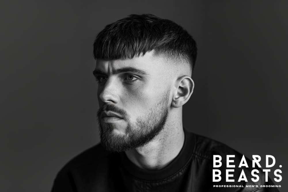 Black and white portrait of a man with a stylish textured haircut and a neatly trimmed beard. His facial hair growth appears deliberate and maintained, but with some signs of slow beard growth. The image highlights a modern grooming aesthetic and professional styling, featuring the Beard Beasts logo in the bottom right corner, emphasizing men's grooming expertise.