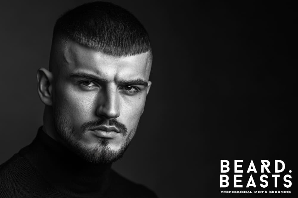 Black and white image of a confident man with neatly trimmed facial hair and a sharp hairstyle, wearing a black turtleneck against a dark background. His facial hair appears light and well-maintained, hinting at the potential challenges of slow beard growth.