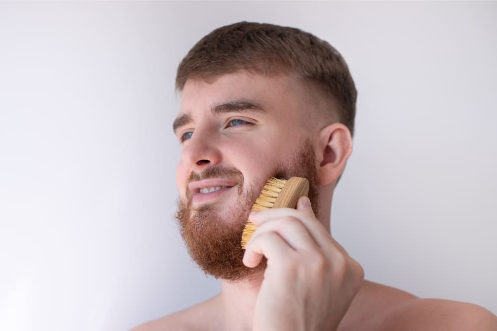 A young man with a short red beard is using a wooden brush to groom his facial hair. He has a relaxed expression and stands against a neutral white background, showcasing a personal grooming routine.