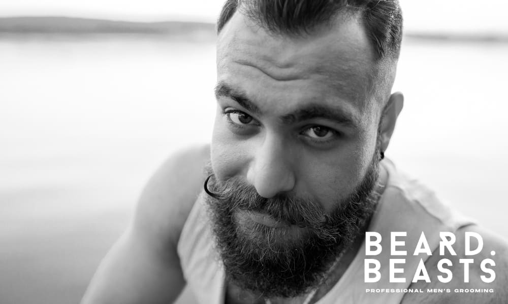 Black and white image of a bearded man with a styled mustache, gazing confidently at the camera by a lake. His well-groomed beard, showcasing a mix of natural texture and control, highlights the challenge of managing beard hair sticking out.