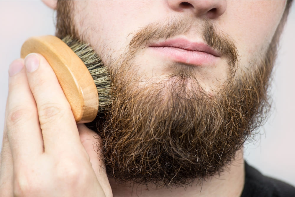 Close-up image of a man grooming his beard using a wooden-handled boar bristle brush. The beard appears thick and slightly frizzy, indicating the need for regular maintenance and care. The act of brushing emphasizes the importance of proper beard grooming for a healthier, tangle-free appearance.