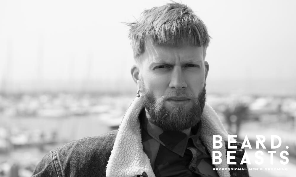 Black and white image of a man with a modern cropped hairstyle and a well-groomed short beard, standing outdoors with a confident expression.