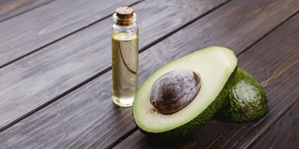 A glass bottle filled with avocado oil for beard care, placed next to a fresh avocado sliced in half, revealing its green flesh and large seed, set on a rustic wooden table.