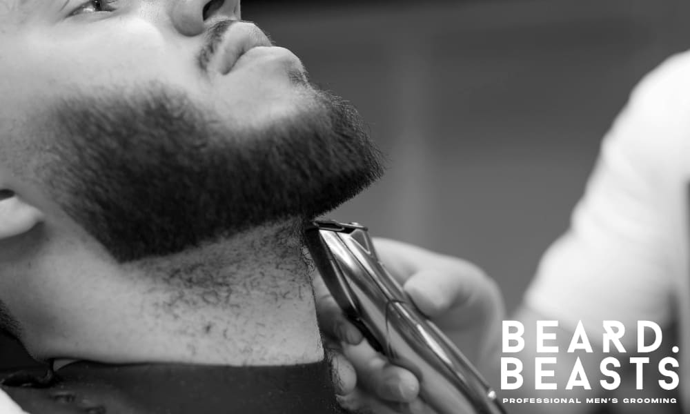 Close-up of a barber trimming a man’s beard neckline with clippers, reinforcing the importance of proper grooming alongside beard growth.