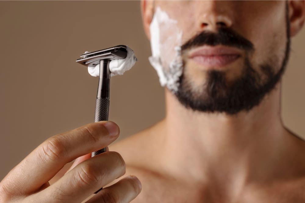 A close-up image of a shirtless man with a well-groomed dark beard, holding a safety razor covered in shaving cream. The man has shaving foam applied to one side of his face, indicating he is in the process of grooming or shaving his facial hair. The focus is on the razor and his hand, while his face is partially visible with a neutral expression. The background is a soft, neutral color, creating a clean and minimalist appearance.