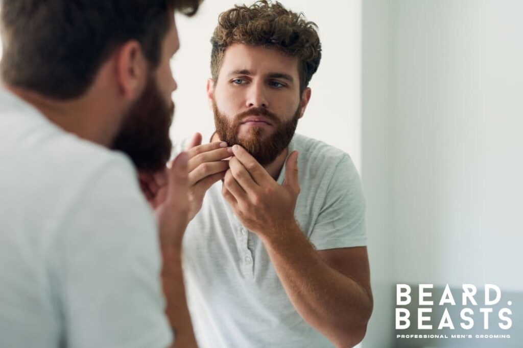A bearded man examining his beard in the mirror, looking concerned about beard acne, with the Beard Beasts logo in the corner.