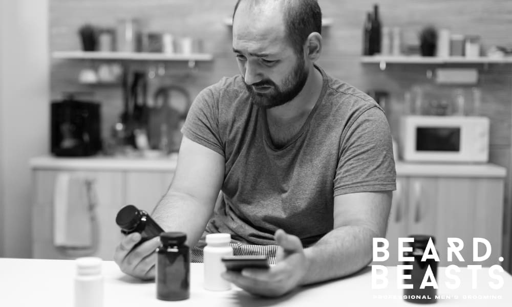 Balding man in a gray t-shirt sitting at a kitchen table, holding supplement bottles and a phone, looking frustrated and unsure.