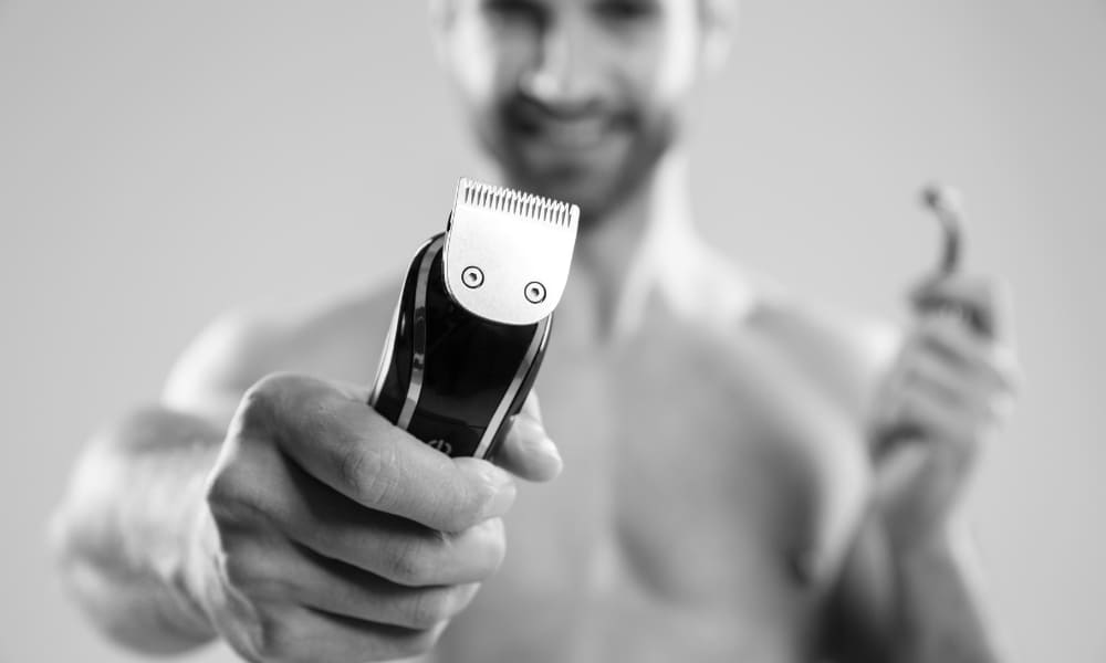 Close-up of a smiling bearded man holding an electric beard trimmer toward the camera, highlighting essential beard grooming tools.