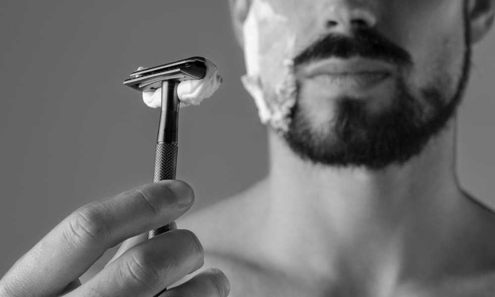 Close-up of a man holding a safety razor with shaving cream on his face, highlighting precise beard line grooming.
