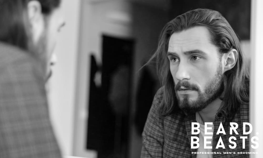 Man with long hair and a trimmed beard looking at his reflection in the mirror, questioning beard growth and grooming results.