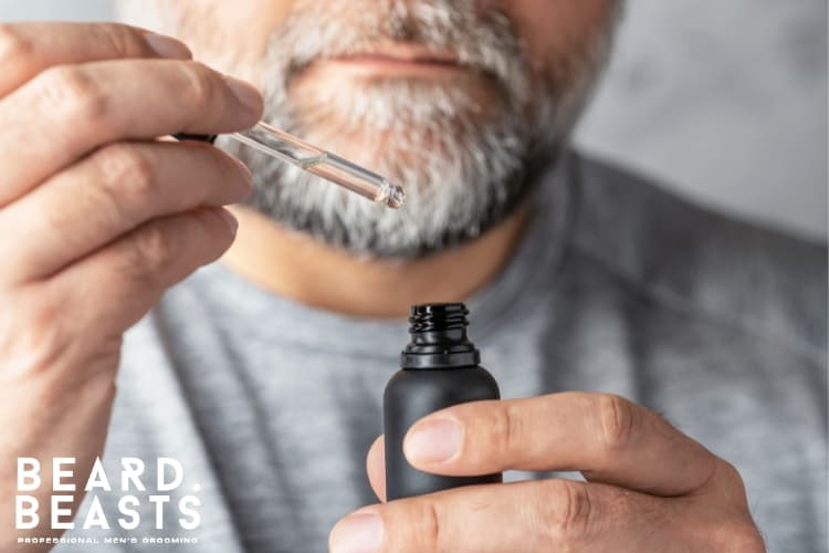 Close-up of a man holding a bottle of beard oil with a dropper, showcasing an essential grooming product for softening and hydrating a rough beard, featuring the Beard Beasts logo in the corner.