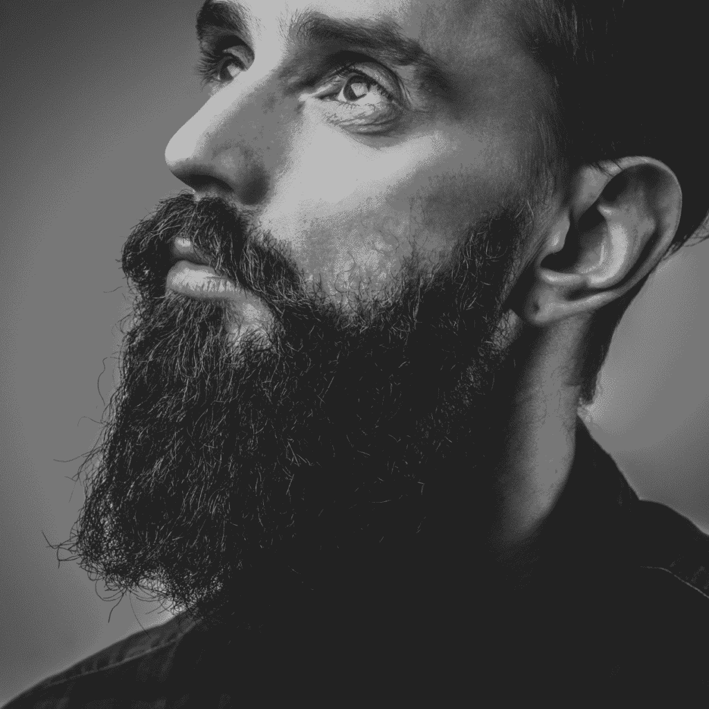 A black and white close-up portrait of a man with a full, frizzy beard. The man gazes thoughtfully upwards, his face illuminated with soft lighting that highlights the texture of his thick, curly beard and the sharpness of his facial features. The background is blurred, keeping the focus on the man's expressive eyes and detailed beard.